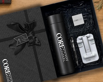 Pode incluir: Uma caixa de presente preta contendo uma garrafa térmica preta, uma caneta preta, um suporte de telefone branco e um adaptador de energia quadrado branco, todos com o logotipo da Core Business Academy. A caixa possui uma fita preta e etiqueta.