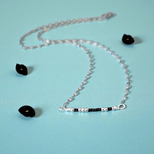 May include: A delicate silver necklace with a bar pendant featuring black and silver beads. The necklace is displayed on a light blue surface, with three small black flower-shaped objects scattered around it.
