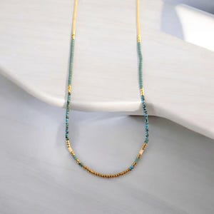 May include: A delicate gold, blue, and white beaded necklace. The necklace is made of small, evenly spaced beads and is displayed on a white surface.