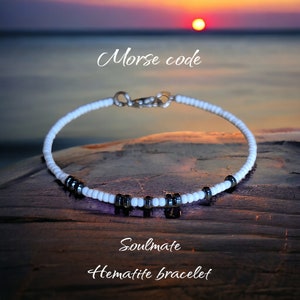 May include: White and black beaded bracelet with the word "Soulmate" written below it. The bracelet is made with white beads and black hematite beads. The text "Morse code" is written above the bracelet.