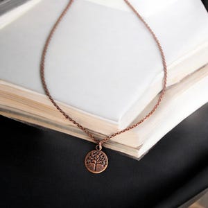 May include: A copper-colored necklace with a tree pendant. The pendant is a small, circular charm with a detailed tree design. The necklace chain is delicate and rests on a stack of open books, with a black surface in the background.