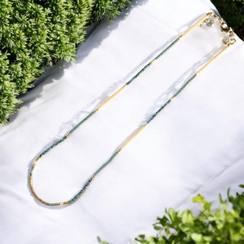May include: A long, delicate necklace made of small, turquoise-colored beads and gold beads. The necklace is on a white background.