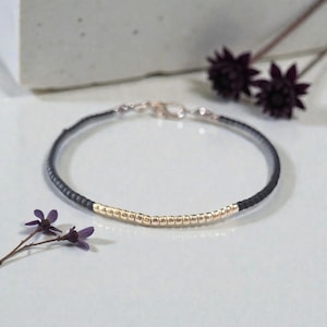 May include: A delicate bracelet featuring a black cord with a section of small, gold-colored beads. The bracelet has a silver clasp and is displayed on a light surface, with dark purple flowers in the background.