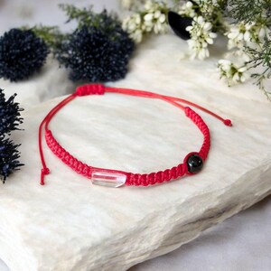 May include: Red braided bracelet with a clear rectangular crystal and a black bead. The bracelet is on a white stone surface, with dark blue and white flowers in the background. The bracelet is adjustable.