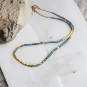 May include: A delicate necklace made with small blue, white, and gold beads. The necklace is layered with two strands of beads.