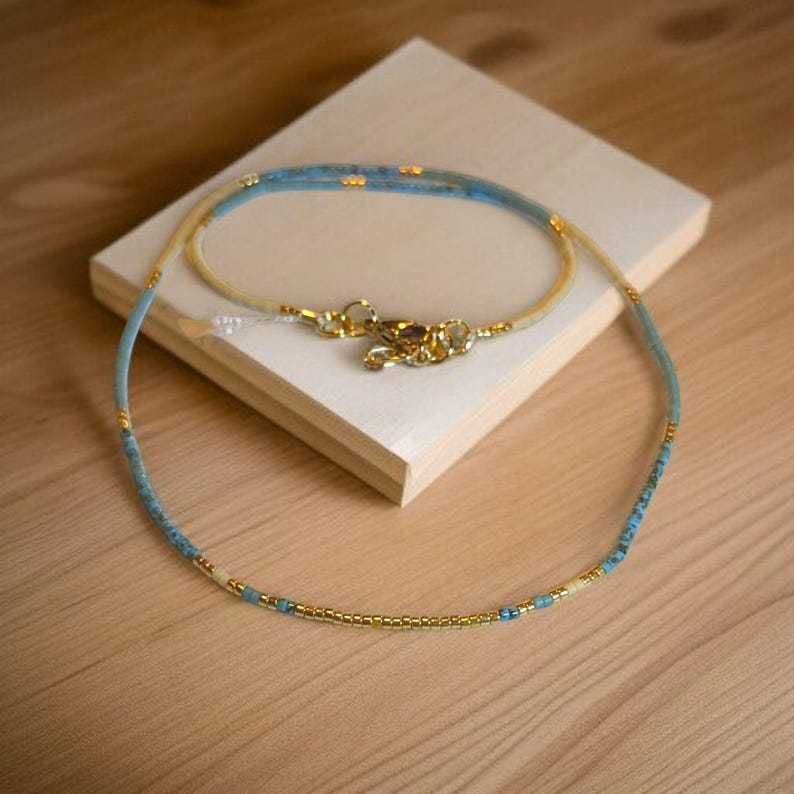 May include: A delicate necklace made with tiny blue and gold beads. The necklace is layered with two strands of beads, creating a subtle ombre effect.