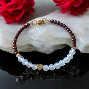 May include: A delicate bracelet with small, round, white beads and gold heart charms. The bracelet is made with dark red beads and a gold clasp.