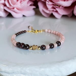 Puede incluir: Un brazalete de cuentas rosa y marrón con un dije de corazón dorado. El brazalete está hecho con pequeñas cuentas redondas y tiene una banda elástica.