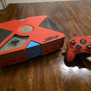 May include: A custom red, blue, and black Xbox video game console featuring JSRF branding and a graphic on the top panel, accompanied by a matching red wired controller with a central Xbox logo and multi-colored action buttons.