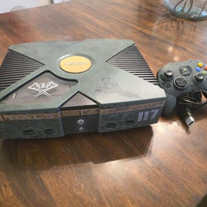 May include: A limited edition Halo themed Xbox video game console featuring a dark green camouflage finish, the number 117, and a matching dark green wired controller. The console has a central circular gold logo and a transparent window section.