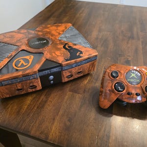 May include: An orange and black Xbox console and controller set. The console features the Xbox logo and a Half-Life 2 logo. The controller has the Xbox logo and multiple colored buttons. The set has a distressed, textured finish.
