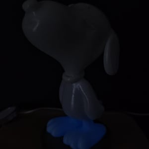 Peut inclure: Une lampe figurine Snoopy avec une tête et un torse blancs dotés d'un collier, éclairée par une douce lumière bleue émanant des pieds. Le personnage est représenté en position debout sur un fond sombre, créant un effet lumineux.
