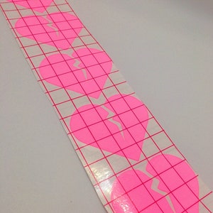 May include: A sheet of pink vinyl stickers with five broken heart designs. The hearts are outlined in white and have a white crack running through them. The stickers are on a gridded backing sheet.