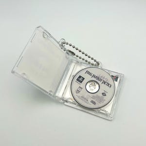 May include: A clear plastic case with a silver chain attached. The case holds a PlayStation game disc with the title "Final Fantasy Tactics" printed on the label.