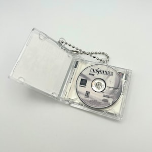 Op de afbeelding: Een transparante plastic sleutelhanger met een zilveren ketting. De sleutelhanger bevat een CD-hoes met een PlayStation-gamedisc erin. De disc is gelabeld met "Final Fantasy IX".