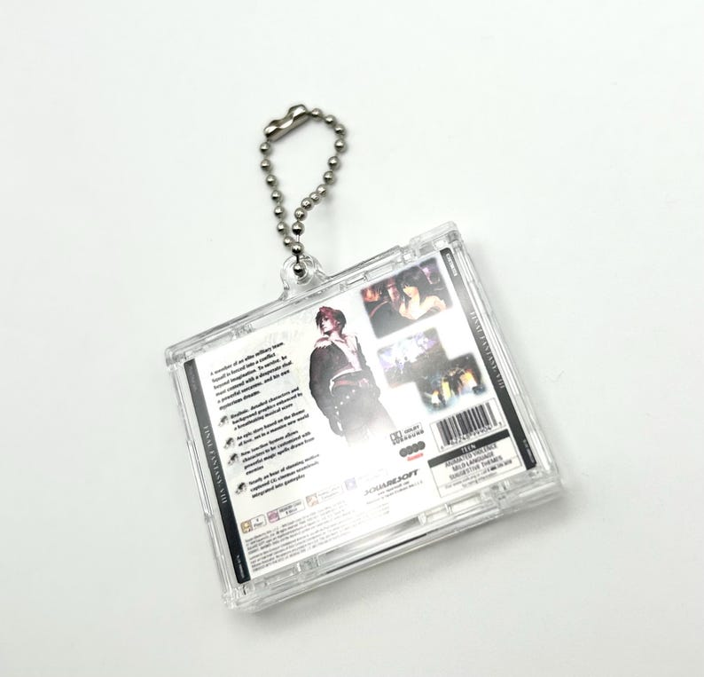 May include: A clear plastic keychain featuring the back of a Final Fantasy VII video game case. The case has a black and white design with a character from the game on the front. The keychain has a silver chain.