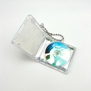 May include: A clear plastic CD case with a silver ball chain attached. The case is open, revealing a CD with a colorful design featuring blue, green, and white hues. The CD case is on a white surface.