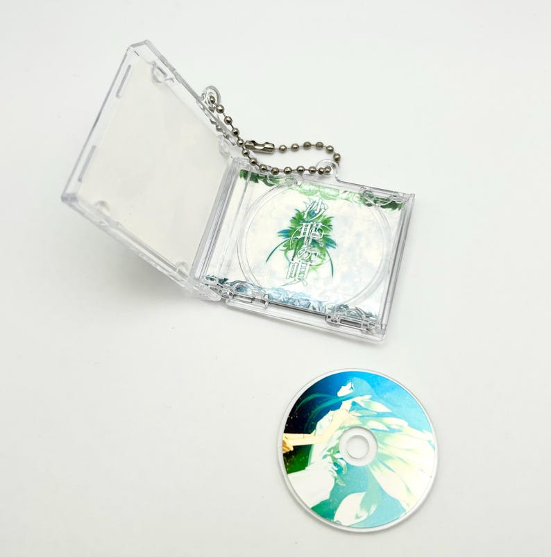 May include: A clear plastic CD case with a silver chain attached, holding a CD with Japanese text and a green and blue design. A loose CD with a similar design lies below.