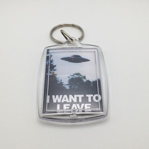 May include: A clear plastic keychain with a black and white image of a UFO and the text "I WANT TO LEAVE".