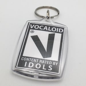 May include: A clear plastic keychain with a black and white logo that says "VOCALOID" with a registered trademark symbol and a large black "V" in the center. The logo also says "CONTENT RATED BY IDOLS".