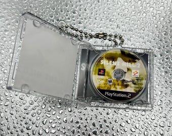 May include: A miniature clear plastic keychain case containing a small circular disc labeled Silent Hill 2 for PlayStation 2. The disc features a yellow and black graphic design with the Konami logo and an M rating symbol for mature audiences.