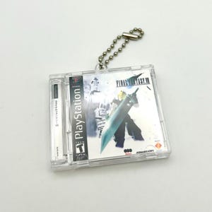 May include: A clear plastic keychain featuring a replica of the Final Fantasy VII video game case. The case is silver and blue with the title "FINAL FANTASY VII" printed on the front. The keychain has a silver chain attached to it.