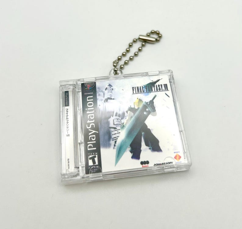 May include: A clear plastic keychain featuring a replica of the Final Fantasy VII video game case. The case is silver and blue with the title "FINAL FANTASY VII" printed on the front. The keychain has a silver chain attached to it.