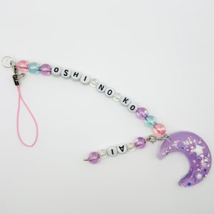 May include: A beaded charm with the text "OSHINO KO" and "AI". The charm features a purple crescent moon pendant with embedded stars and glitter. The beads are in pastel colors, and the charm has a pink lanyard.