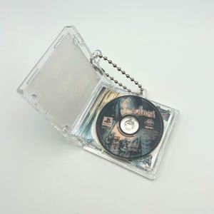 May include: A clear plastic keychain with a silver chain. The keychain opens to reveal a video game disc with a woman on the cover. The disc says "Koudelka" and "Disc 1".