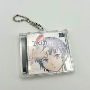 May include: A clear plastic keychain with a silver chain. The keychain features a CD case with a picture of a young girl with purple hair. The text "lain" is printed on the CD case.