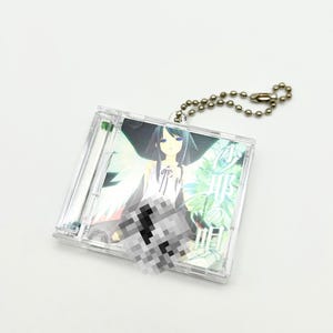 May include: A clear plastic CD case keychain featuring an anime-style illustration of a woman with long dark hair and blue eyes. The artwork has a fantasy theme with green and white accents. A silver ball chain is attached for hanging.