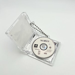 Könnte beinhalten: Ein transparentes Plastikgehäuse mit einer silbernen Kette, in dem sich eine Final Fantasy VII Videospiel-Disc befindet. Die Disc ist mit "FINAL FANTASY VII" beschriftet und trägt das PlayStation-Logo.