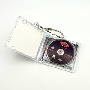 May include: A clear plastic keychain case containing a Resident Evil PlayStation game disc. The disc is black with red and white text, and the case is attached to a silver ball chain.
