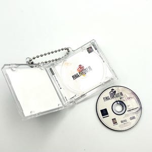 May include: A clear plastic case with a silver chain, holding a video game disc with the text "Final Fantasy VII" on it. The disc is in a clear plastic case with a silver chain. The case is open and the disc is visible. There is a second disc on the table, also with the text "Final Fantasy VII" on it.