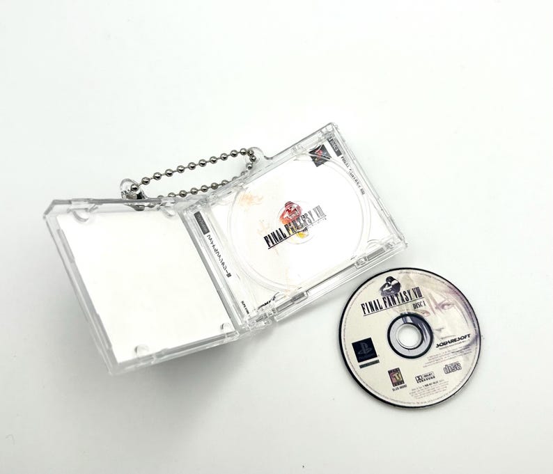 May include: A clear plastic case with a silver chain, holding a video game disc with the text "Final Fantasy VII" on it. The disc is in a clear plastic case with a silver chain. The case is open and the disc is visible. There is a second disc on the table, also with the text "Final Fantasy VII" on it.