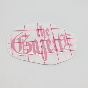 May include: Pink vinyl sticker with the text "The Gazette" in a gothic font.
