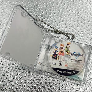 May include: A miniature clear plastic DVD case containing a Kingdom Hearts game disc for PlayStation 2, featuring illustrations of Sora, Donald Duck, and Goofy. The case is attached to a silver metal ball chain for use as a keychain accessory.