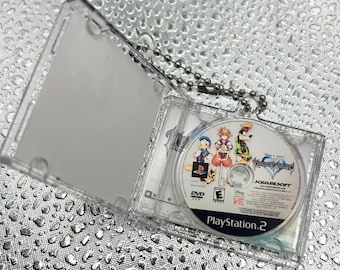 May include: A miniature clear plastic DVD case containing a Kingdom Hearts game disc for PlayStation 2, featuring illustrations of Sora, Donald Duck, and Goofy. The case is attached to a silver metal ball chain for use as a keychain accessory.