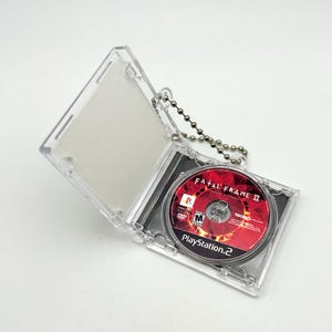 Op de afbeelding: Een doorzichtige plastic hoes met een zilveren kogelketting eraan, met een PlayStation 2-game disc. De disc heeft een rood en zwart ontwerp met de titel "FATAL FRAME II" en het PlayStation 2-logo.