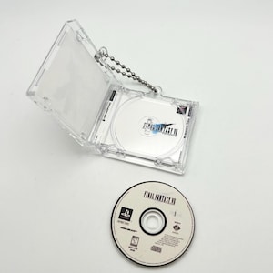 May include: A clear plastic case with a silver chain attached to it. The case holds a video game disc with the text "FINAL FANTASY VII" on it. The disc is also visible outside of the case.