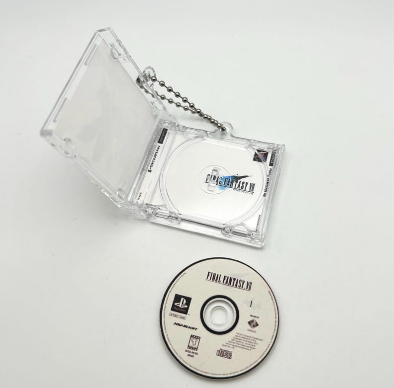 May include: A clear plastic case with a silver chain attached to it. The case holds a video game disc with the text "FINAL FANTASY VII" on it. The disc is also visible outside of the case.