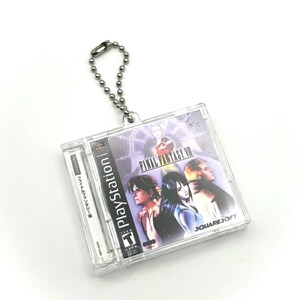 May include: A clear plastic keychain featuring a replica of the Final Fantasy VIII PlayStation game case. The case has a purple and blue background with a picture of the main characters and the text "FINAL FANTASY VIII" and "SQUARE SOFT".
