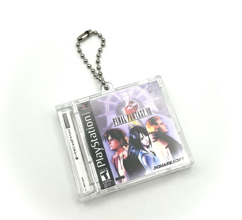 May include: A clear plastic keychain featuring a replica of the Final Fantasy VIII PlayStation game case. The case has a purple and blue background with a picture of the main characters and the text "FINAL FANTASY VIII" and "SQUARE SOFT".