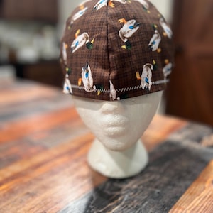 May include: A brown cap with a repeating pattern of white, blue, and orange ducks. The cap is displayed on a white mannequin head. The background is a wooden table.