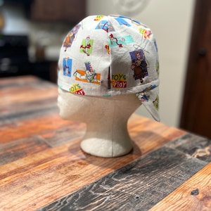 May include: A white scrub cap featuring a colorful repeating pattern of various cartoon characters from the Toy Story franchise, including Woody, Buzz Lightyear, and Forky, along with the text Toys at Play printed in red and green lettering.