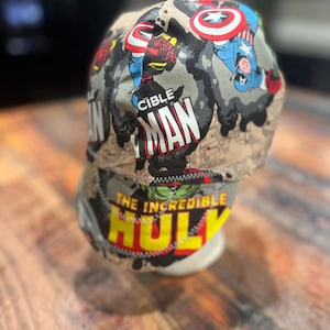 May include: A beige and gray comic book-themed cap featuring Captain America, Spider-Man, and The Incredible Hulk. The cap has the words "Cible Man" and "The Incredible Hulk" printed on it. The cap is displayed on a white form.
