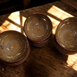 May include: A collection of four ceramic bowls with a speckled, glazed interior and a dark brown rim. The bowls are arranged on a wooden surface, with natural light casting shadows across the scene. The bowls have a rustic, handcrafted appearance.