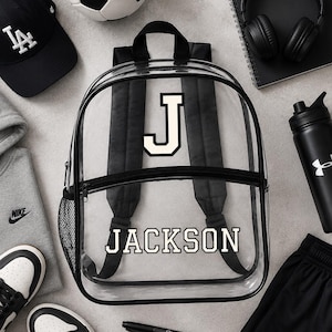 May include: A transparent backpack with the name "JACKSON" in white lettering, a black baseball cap, a grey hoodie, black and white trainers, a football, black shorts, a water bottle, and black headphones. A black Sharpie pen is also visible.