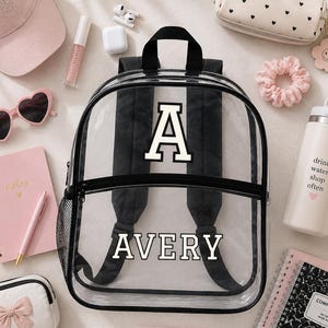Puede incluir: Una mochila transparente con correas negras y el nombre "AVERY" en letras blancas. La mochila está rodeada de accesorios rosas, incluyendo un cuaderno, gafas de sol, una botella de agua y un estuche. También se ven una gorra de béisbol rosa y una goma para el pelo.