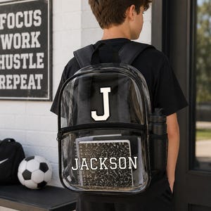 Puede incluir: Una mochila transparente con ribete negro y una 'J' blanca. El nombre 'JACKSON' está impreso en un cuaderno en el interior. Una botella de agua negra está en el bolsillo lateral. Una bolsa de deporte negra y un balón de fútbol están sobre una mesa.